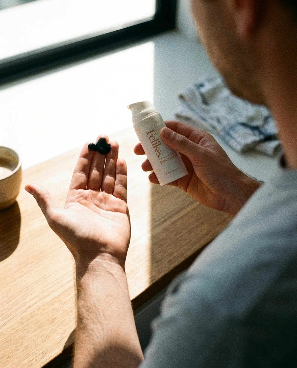A dose of Feliks The Exfoliator charcoal gel on a man's fingertips at a timber kitchen bench, bottle in his other hand, morning window light