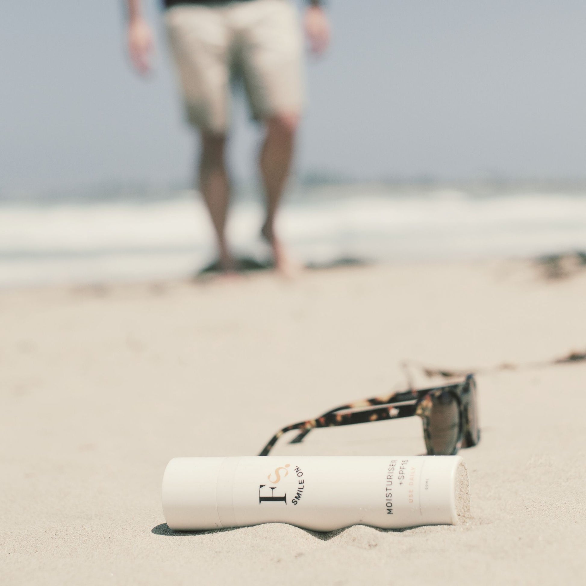 Feliks daily moisturiser and sunglasses on sand with blurred person in background