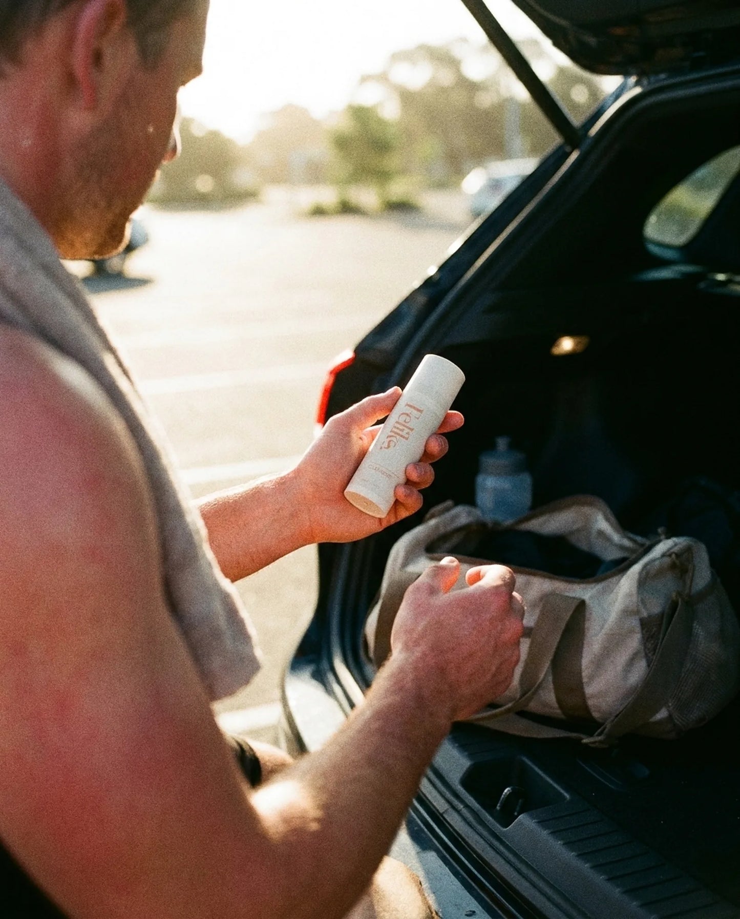 A man at the open boot of his car after training, towel round his neck, holding Feliks The Cleanser he has just taken from his gym bag in early morning light